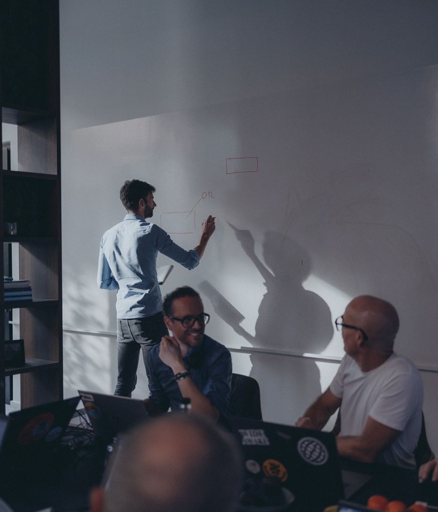 ONE-Consultants team working together on a problem. One person is writing a schema on a whiteboard, while two other men are sitting at a desk behind their computers